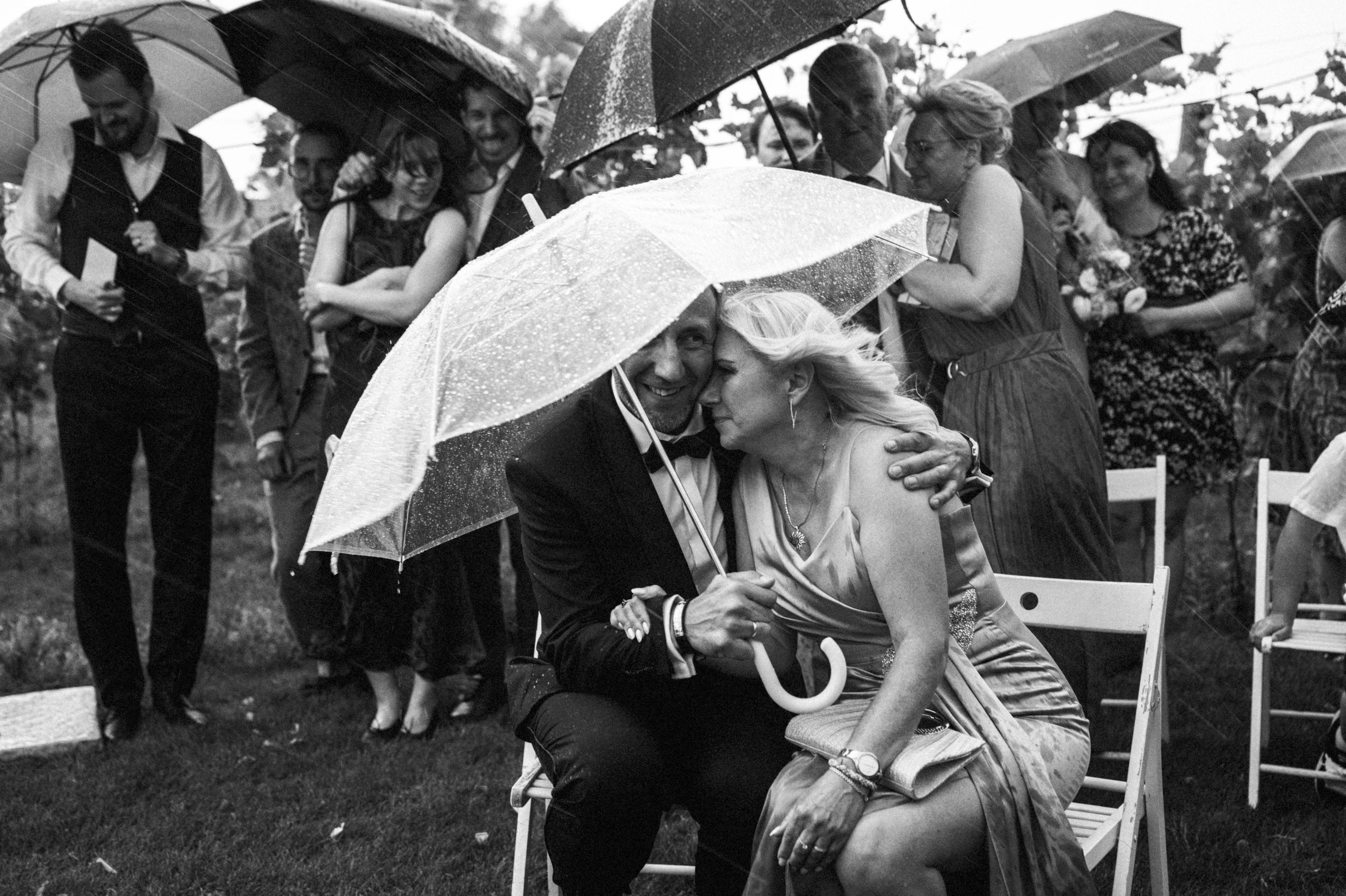 Goście przytulają się pod parasolem podczas deszczowej ceremonii. Czarno-biała fotografia.